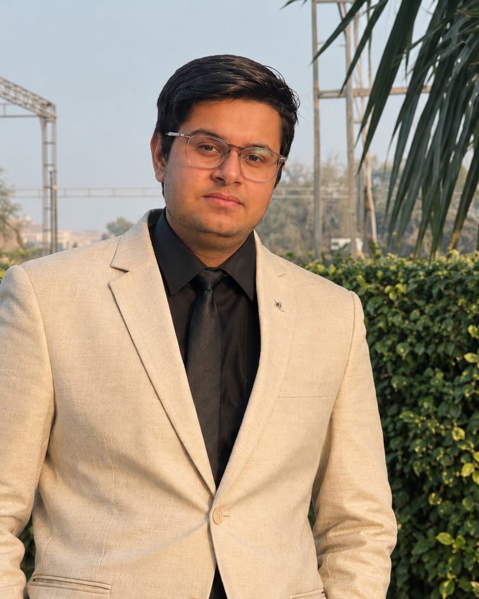 Sanat B Bajaj in a cream blazer, black shirt and black tie, standing outdoors by a hedge